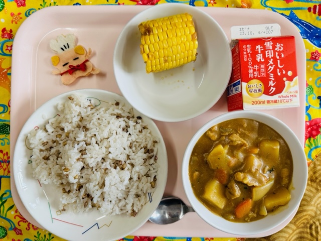 ピンクのトレーに給食が右上から反時計回りで牛乳、やきとうもろこし、コック帽をかぶった布製マスコット、ごはん、カレーと並べられている写真