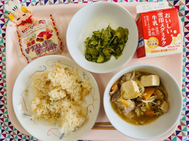 ピンクのトレーに給食が右上から反時計回りで牛乳、ピリからきゅうり、さつまいもと栗のタルト、コック帽をかぶった布製マスコット、かしわずし、にくどうふと並べられている写真