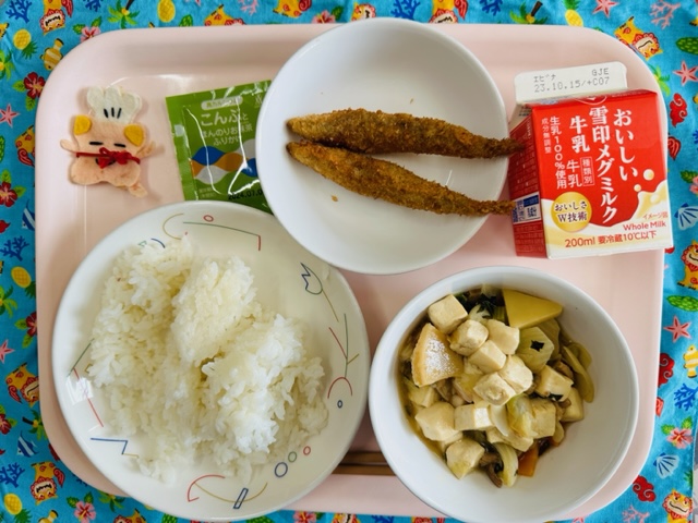 ピンクのトレーに給食が右上から反時計回りで牛乳、ししゃもフライ、ふりかけ、コック帽をかぶった布製マスコット、ごはん、豆腐の中華煮と並べられている写真