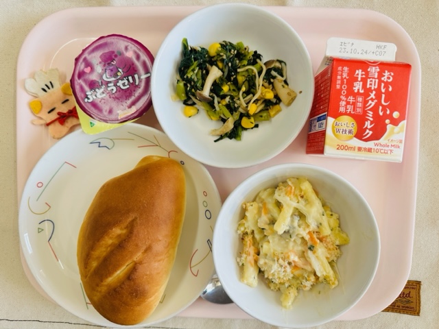 ピンクのトレーに給食が右上から反時計回りで牛乳、小松菜のサラダ、ぶどうゼリー、コック帽をかぶった布製マスコット、ミルクパン、マカロニグラタンと並べられている写真