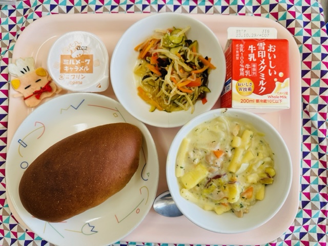 ピンクのトレーに給食が右上から反時計回りで牛乳、しらたきサラダ、デザート、コック帽をかぶった布製マスコット、くろパン、さつまいものクリーム煮と並べられている写真