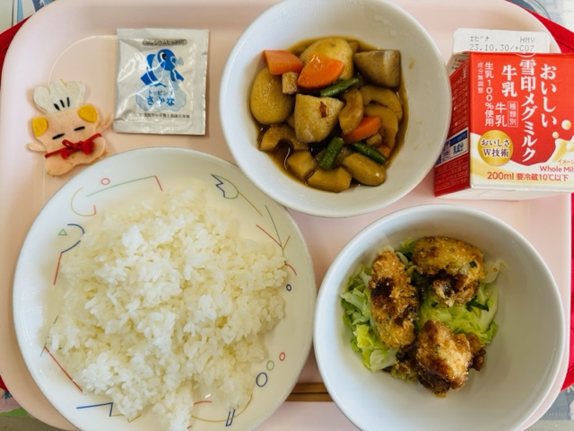 ピンクのトレーに給食が右上から反時計回りで牛乳、さといもの煮物、ふりかけ、コック帽をかぶった布製マスコット、ごはん、チキンかつと並べられている写真