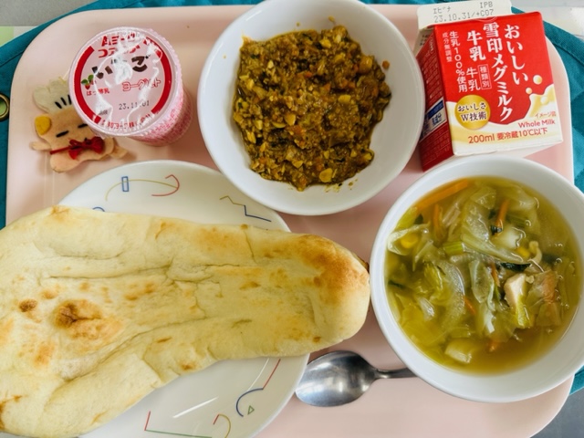 ピンクのトレーに給食が右上から反時計回りで牛乳、ドライカレー、デザート、コック帽をかぶった布製マスコット、ナン、やさいスープと並べられている写真