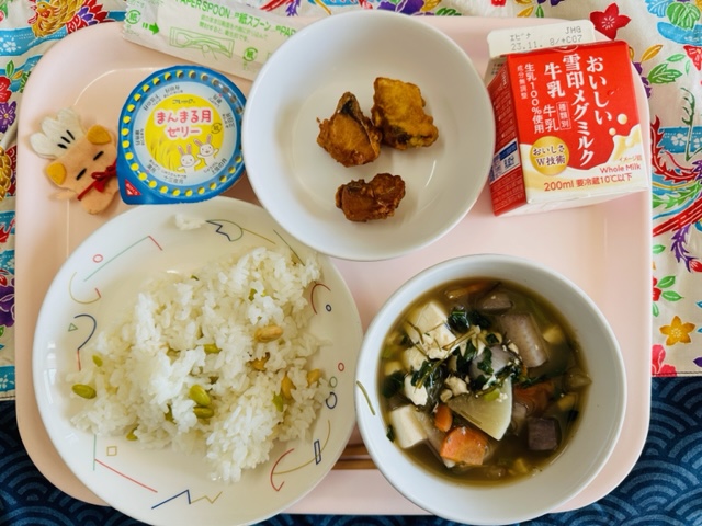 ピンクのトレーに給食が右上から反時計回りで牛乳、さばのカレー揚げ、まんまる月ゼリー、コック帽をかぶった布製マスコット、まめまめごはん、つきみ汁と並べられている写真