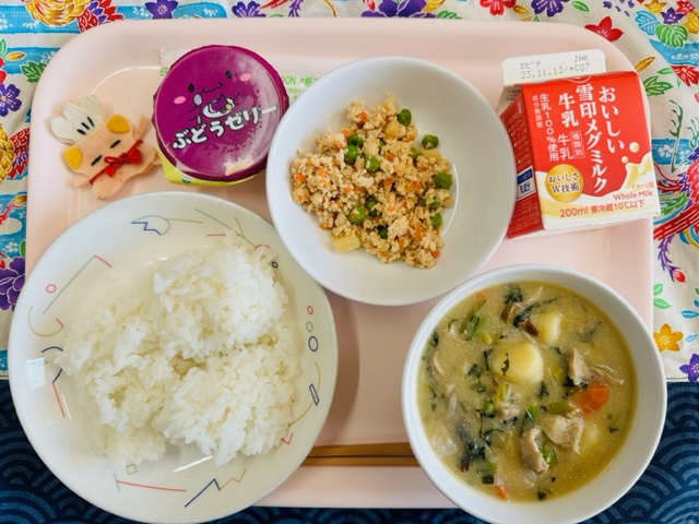 ピンクのトレーに給食が右上から反時計回りで牛乳、とりそぼろ、ぶどうゼリー、コック帽をかぶった布製マスコット、ごはん、いももちじると並べられている写真