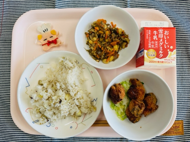 ピンクのトレーに給食が右上から反時計回りで牛乳、五目豆、コック帽をかぶった布製マスコット、さつまいもごはん、さけフライとキャベツソテーと並べられている写真