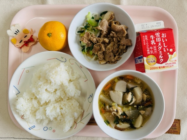 ピンクのトレーに給食が右上から反時計回りで牛乳、豚肉の生姜焼き 野菜炒め添え、みかん、コック帽をかぶった布製マスコット、ごはん、のっぺい汁と並べられている写真