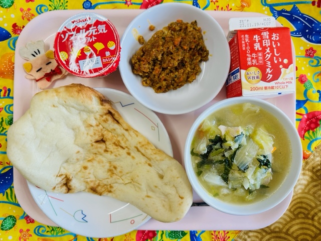 ピンクのトレーに給食が右上から反時計回りで牛乳、キーマカレー、ヨーグルト、コック帽をかぶった布製マスコット、ナン、野菜のスープ煮と並べられている写真