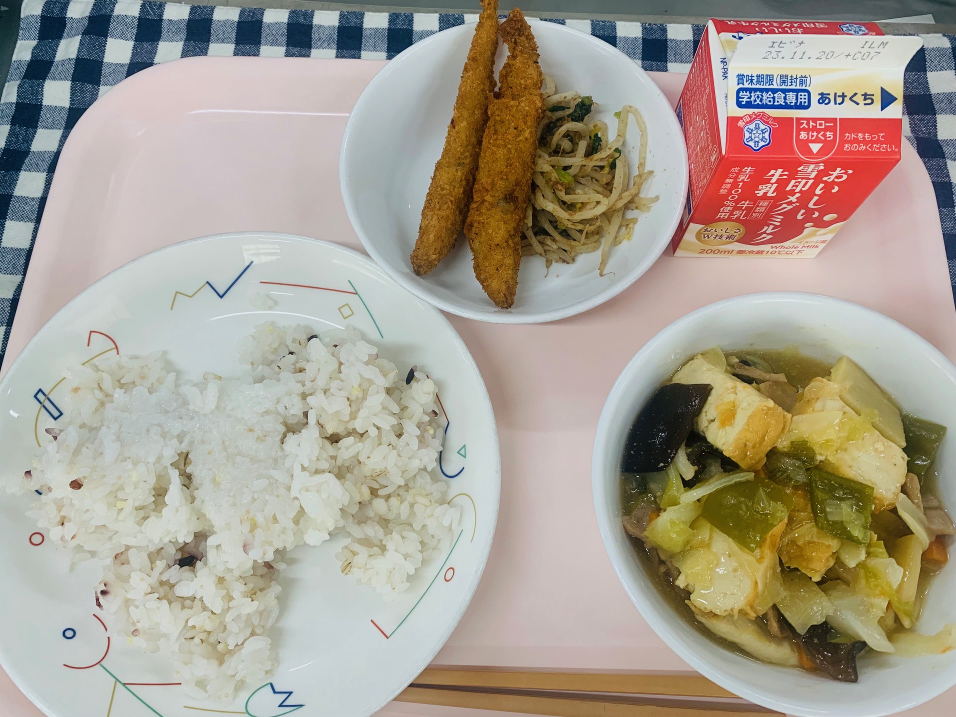 ピンクのトレーに給食が右上から反時計回りで牛乳、ししゃもフライともやしのおかかあえ、十穀米ご飯、ジャージャンどうふと並べられている写真