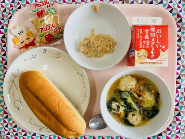 ピンクのトレーに給食が右上から反時計回りで牛乳、ツナサンドの具、アップルパイ、コック帽をかぶった布製マスコット、パン、白菜と肉だんごのスープと並べられている写真