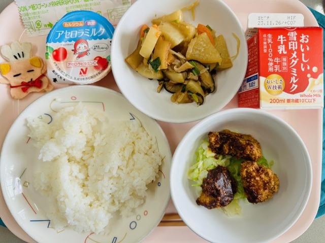 ピンクのトレーに給食が右上から反時計回りで牛乳、大根の炒め煮、アセロラゼリー、コック帽をかぶった布製マスコット、ごはん、みそカツとキャベツソテーと並べられている写真