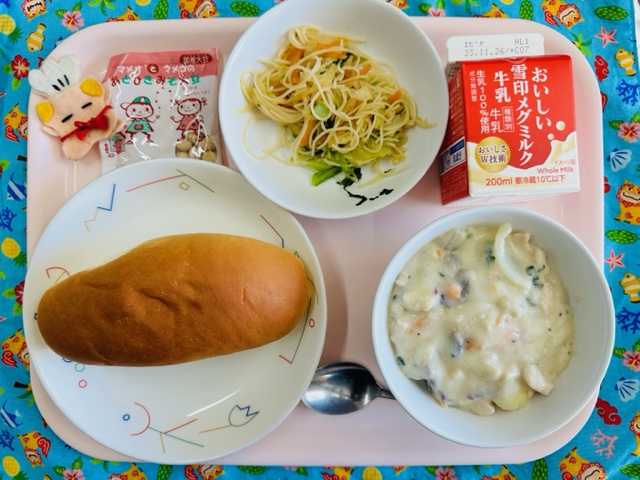 ピンクのトレーに給食が右上から反時計回りで牛乳、ビーフンソテー、みそだいず、コック帽をかぶった布製マスコット、あじつけパン、秋の味覚シチューと並べられている写真