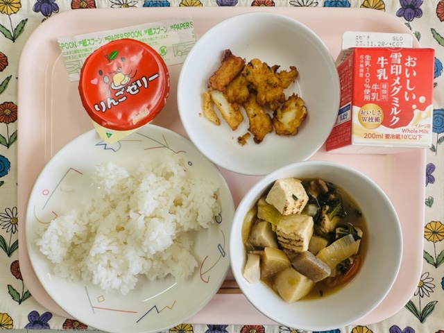 ピンクのトレーに給食が右上から反時計回りで牛乳、いかのふわっとあげ、りんごゼリー、ごはん、里芋と焼き豆腐のうま煮と並べられている写真