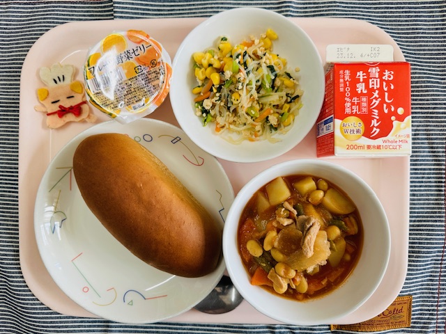 ピンクのトレーに給食が右上から反時計回りで牛乳、しらたきソテー、野菜ゼリー、コック帽をかぶった布製マスコット、くろパン、ポークビーンズと並べられている写真