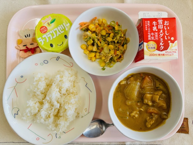 ピンクのトレーに給食が右上から反時計回りで牛乳、ツナコーンサラダ、ラ・フランスゼリー、コック帽をかぶった布製マスコット、ごはん、チキンカレーと並べられている写真