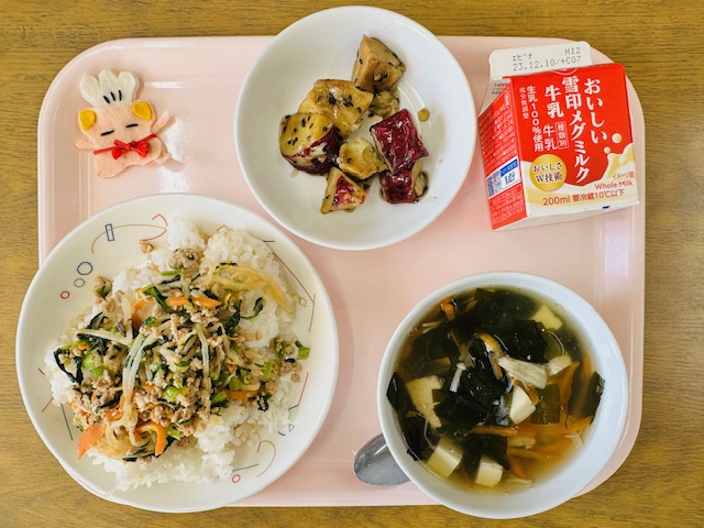 ピンクのトレーに給食が右上から反時計回りで牛乳、大学いも、コック帽をかぶった布製マスコット、ビビンパ、わかめスープと並べられている写真