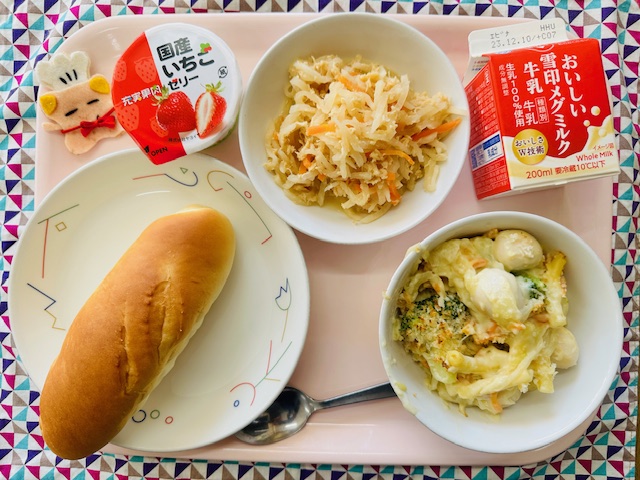 ピンクのトレーに給食が右上から反時計回りで牛乳、だいこんのサラダ、国産いちごゼリー、コック帽をかぶった布製マスコット、ミルクパン、うずらの卵とブロッコリーのグラタンと並べられている写真