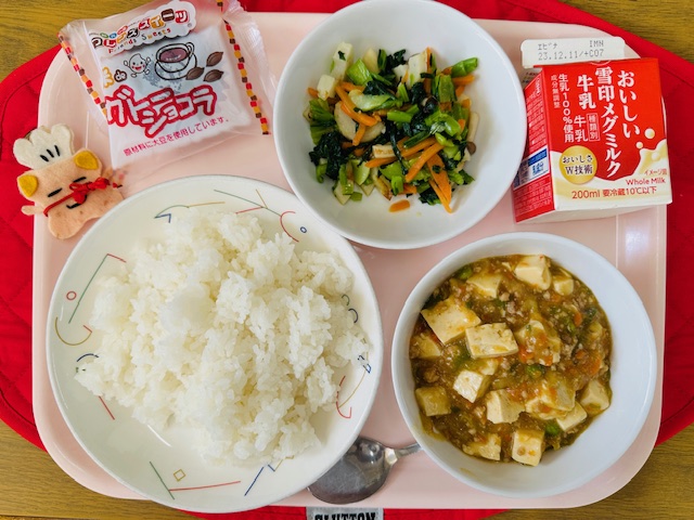 ピンクのトレーに給食が右上から反時計回りで牛乳、青菜炒め、ガトーショコラ、コック帽をかぶった布製マスコット、ごはん、マーボーどうふと並べられている写真