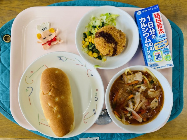 ピンクのトレーに給食が右上から反時計回りでのむヨーグルト、カレーコロッケとミックスソテー、コック帽をかぶった布製マスコット、げんまいパン、イタリアンスープと並べられている写真