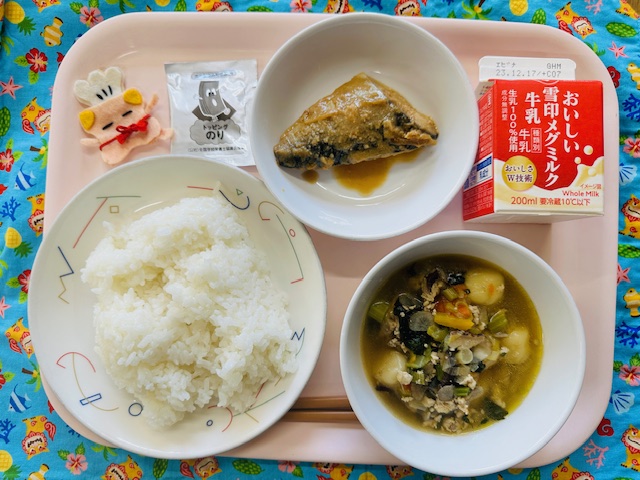 ピンクのトレーに給食が右上から反時計回りで牛乳、さばの味噌煮、ふりかけ、コック帽をかぶった布製マスコット、ごはん、いももちじると並べられている写真