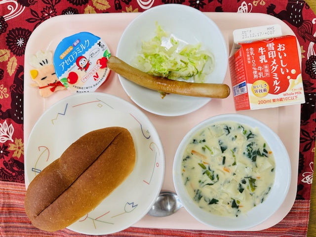 ピンクのトレーに給食が右上から反時計回りで牛乳、ウインナーと野菜、アセロラゼリー、コック帽をかぶった布製マスコット、パン、ほうれん草のミルクスープと並べられている写真
