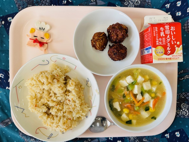 ピンクのトレーに給食が右上から反時計回りで牛乳、ごまみそボール、コック帽をかぶった布製マスコット、ガーリックピラフ、コーン中華スープと並べられている写真