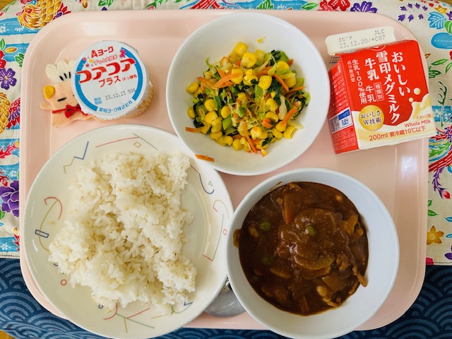 ピンクのトレーに給食が右上から反時計回りで牛乳、野菜ソテー、ヨーグルト、コック帽をかぶった布製マスコット、ごはん、ハヤシライスと並べられている写真