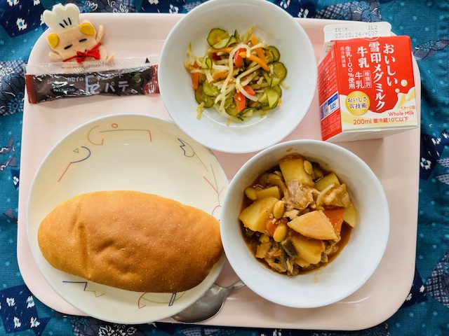 ピンクのトレーに給食が右上から反時計回りで牛乳、切り干し大根のサラダ、チョコパテ、コック帽をかぶった布製マスコット、コッペパン、ポークビーンズと並べられている写真