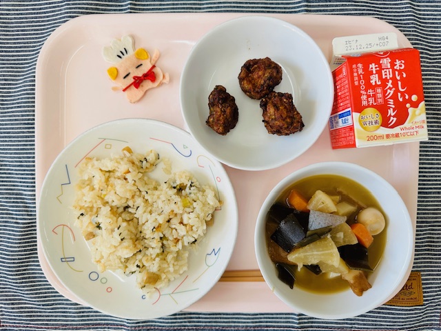 ピンクのトレーに給食が右上から反時計回りで牛乳、豚肉の風味揚げ、コック帽をかぶった布製マスコット、とりなめし、おでんと並べられている写真