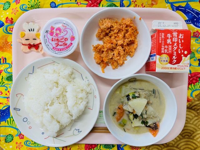 ピンクのトレーに給食が右上から反時計回りで牛乳、さけそぼろ、いちごのフール、コック帽をかぶった布製マスコット、ごはん、ごじると並べられている写真