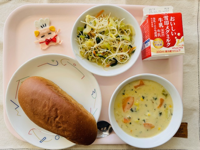 ピンクのトレーに給食が右上から反時計回りで牛乳、ビーフンソテー、コック帽をかぶった布製マスコット、くろパン、白菜とコーンのクリームシチューと並べられている写真