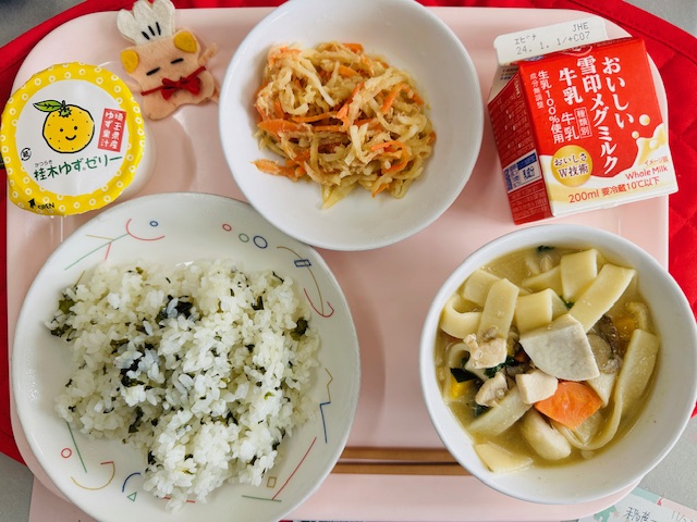 ピンクのトレーに給食が右上から反時計回りで牛乳、だいこんサラダ、かつらきゆずデザート 、コック帽をかぶった布製マスコット、わかめごはん、ほうとうと並べられている写真