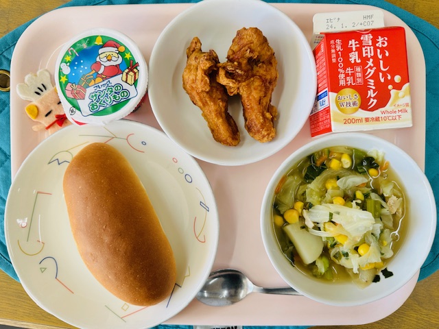 ピンクのトレーに給食が右上から反時計回りで牛乳、フライドチキン、デザート 、コック帽をかぶった布製マスコット、あじつけパン、やさいスープと並べられている写真