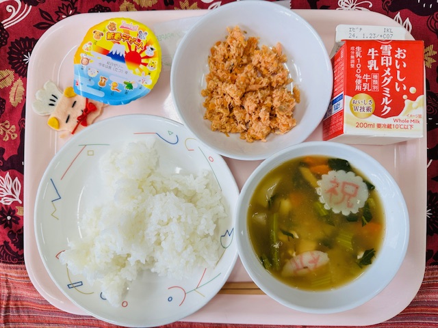 ピンクのトレーに給食が右上から反時計回りで牛乳、さけそぼろ、新春デザート 紅白ゼリー、コック帽をかぶった布製マスコット、ごはん、しらたま雑煮と並べられている写真