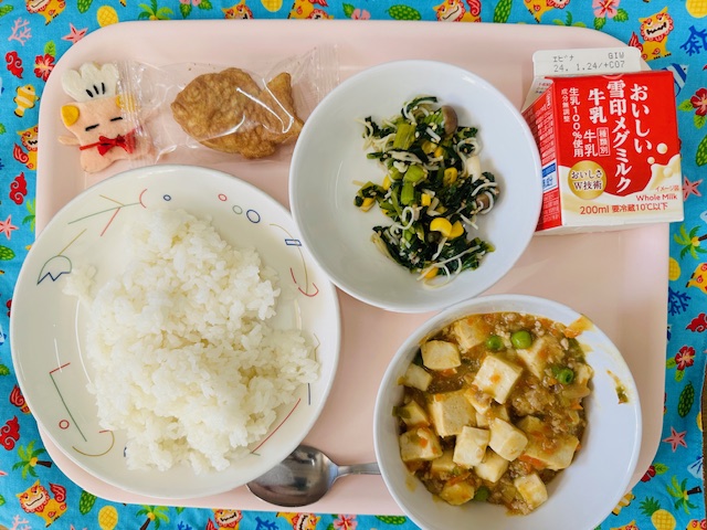 ピンクのトレーに給食が右上から反時計回りで牛乳、小松菜のサラダ、ミニたい焼き、コック帽をかぶった布製マスコット、ごはん、マーボー豆腐と並べられている写真