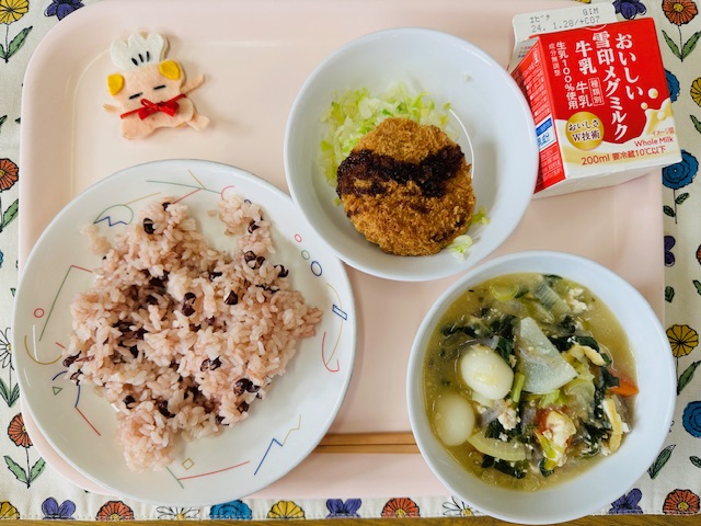 ピンクのトレーに給食が右上から反時計回りで牛乳、メンチカツとキャベツソテー、コック帽をかぶった布製マスコット、あずきごはん、だんごじると並べられている写真