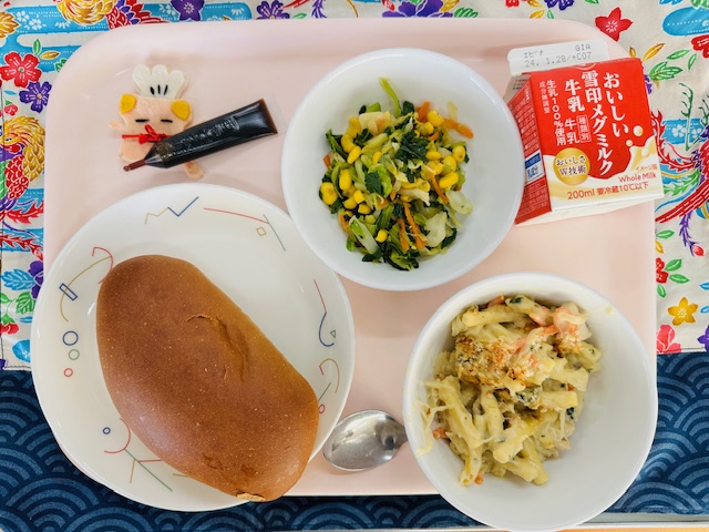 ピンクのトレーに給食が右上から反時計回りで牛乳、やさいソテー、ミルメーク、コック帽をかぶった布製マスコット、くろパン、マカロニグラタンと並べられている写真