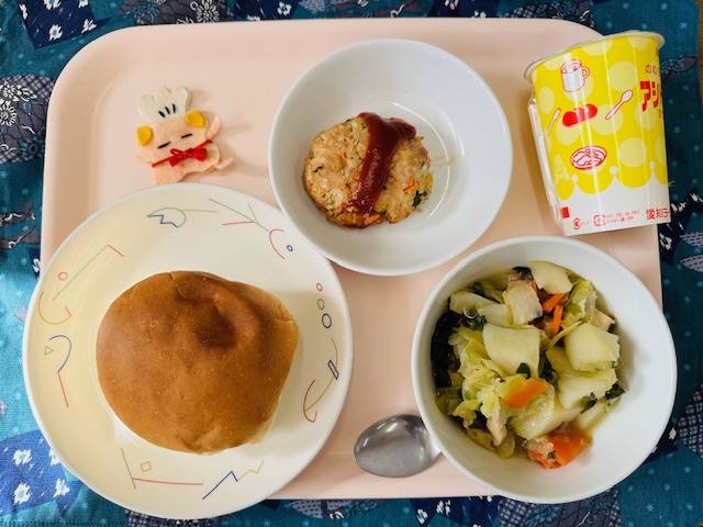 ピンクのトレーに給食が右上から反時計回りで発酵乳、ビーンズハンバーグ、コック帽をかぶった布製マスコット、バーガーパン、やさいのスープにと並べられている写真