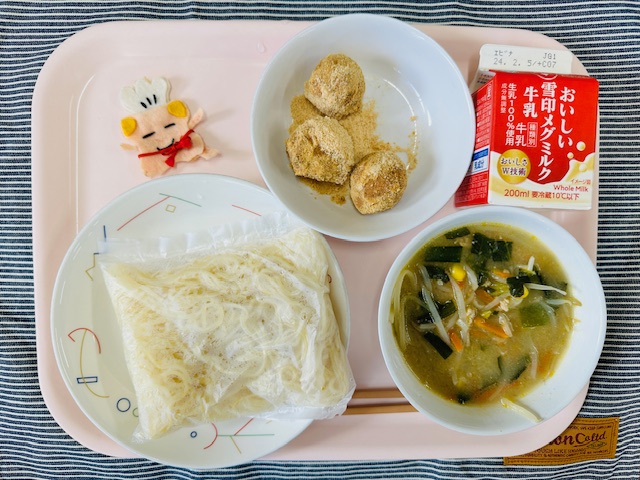 ピンクのトレーに給食が右上から反時計回りで牛乳、きなこドーナツ、コック帽をかぶった布製マスコット、麺、みそラーメンのスープと具と並べられている写真