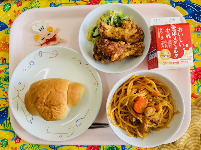 ピンクのトレーに給食が右上から反時計回りで牛乳、フライドチキンとミックスソテー、コック帽をかぶった布製マスコット、ロールパン、スパゲッティナポリタンと並べられている写真