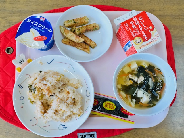 ピンクのトレーに給食が右上から反時計回りで牛乳、サラダのりとフライドポテト、アイスクリーム、コック帽をかぶった布製マスコット、キムチごはん、わかめスープと並べられている写真