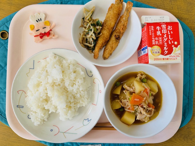 ピンクのトレーに給食が右上から反時計回りで牛乳、ししゃもフライともやしのおかかあえ、コック帽をかぶった布製マスコット、ごはん、肉じゃがと並べられている写真