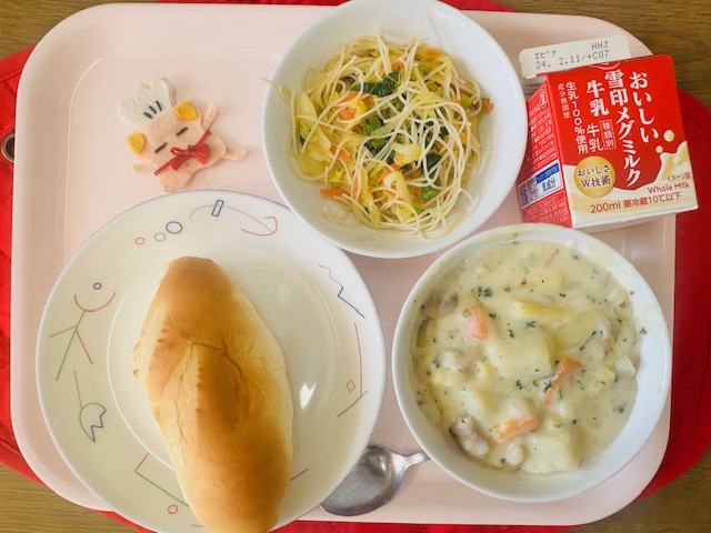 ピンクのトレーに給食が右上から反時計回りで牛乳、ビーフンソテー、コック帽をかぶった布製マスコット、ミルクパン、とりにくのクリーム煮と並べられている写真