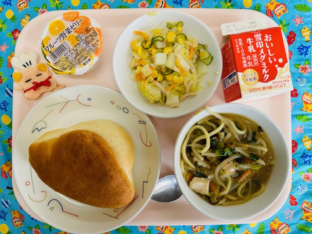 ピンクのトレーに給食が右上から反時計回りで牛乳、ポテサラ、フルーツ野菜ゼリー、コック帽をかぶった布製マスコット、パン、ちゃんぽんスープと並べられている写真