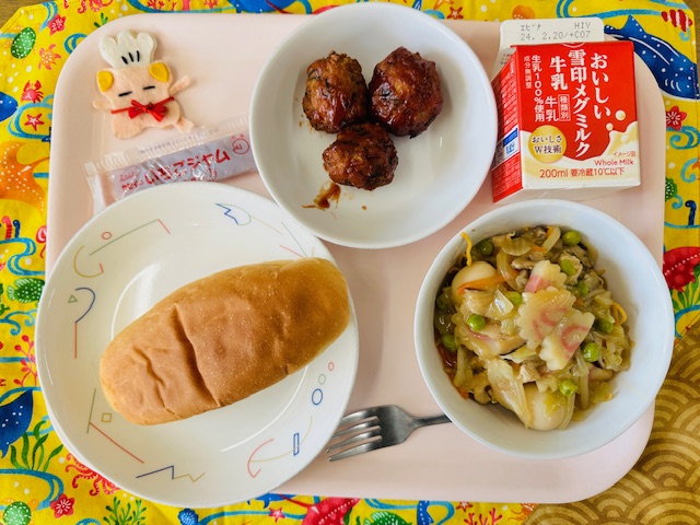 ピンクのトレーに給食が右上から反時計回りで牛乳、とうふボールのケチャップソースあえ、いちごジャム、コック帽をかぶった布製マスコット、コッペパン、やきそばあんかけと並べられている写真