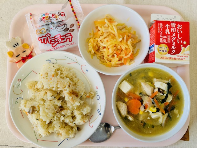 ピンクのトレーに給食が右上から反時計回りで牛乳、だいこんサラダ、ガトーショコラ、コック帽をかぶった布製マスコット、チキンピラフ、コーン中華スープと並べられている写真