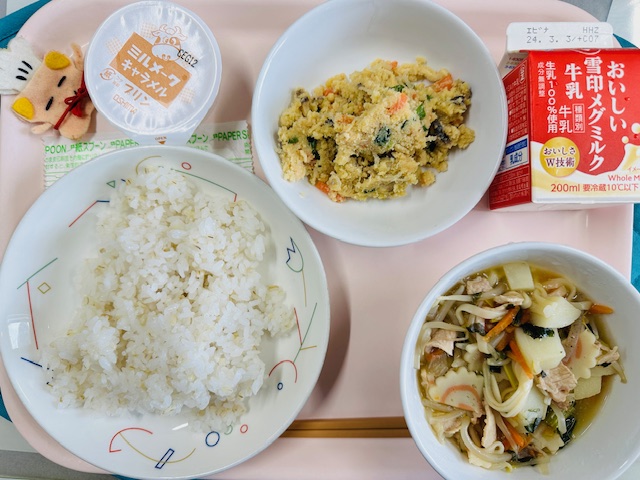 ピンクのトレーに給食が右上から反時計回りで牛乳、うのはな、ミルメークキャラメル、コック帽をかぶった布製マスコット、ごはん、あんかけじると並べられている写真