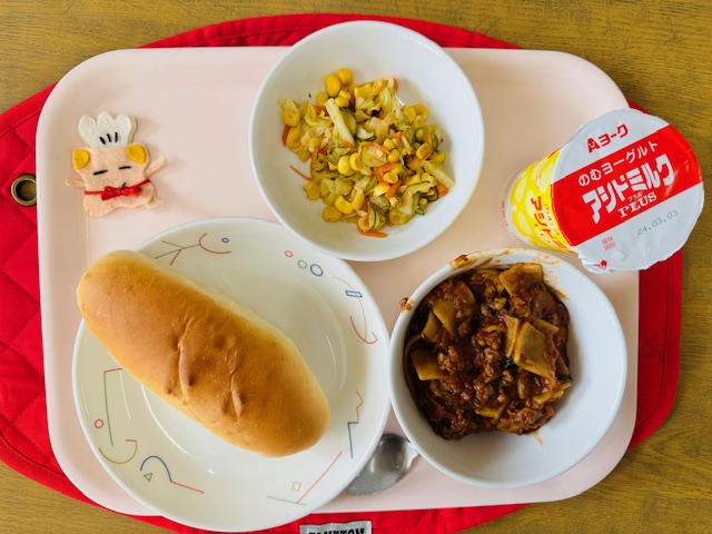 ピンクのトレーに給食が右上から反時計回りでのむヨーグルト、コーンサラダ、コック帽をかぶった布製マスコット、ミルクパン、ラザニアと並べられている写真