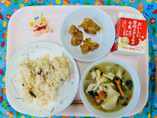 ピンクのトレーに給食が右上から反時計回りで牛乳、鶏肉のレモン醤油和え、コック帽をかぶった布製マスコット、かやくごはん、すいとんと並べられている写真