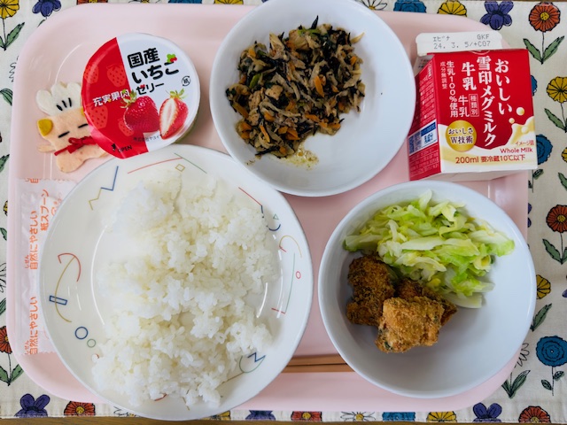 ピンクのトレーに給食が右上から反時計回りで牛乳、切り干し大根のミルク煮、国産いちごゼリー、コック帽をかぶった布製マスコット、ごはん、さけフライとキャベツソテーと並べられている写真